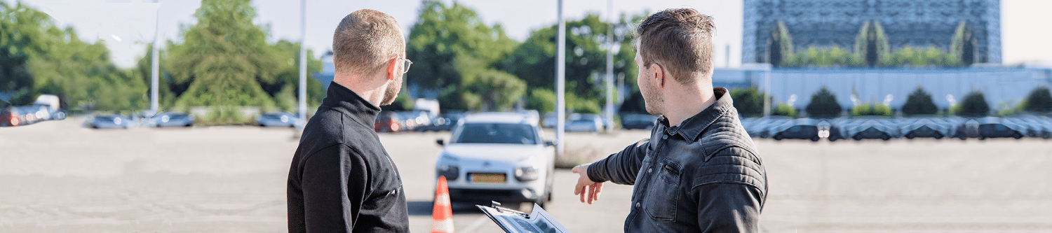 2 men looking at a parked car behind a traffic cone in a training lot