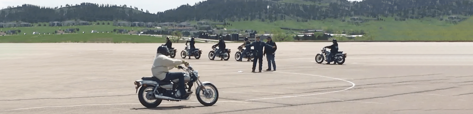 motorcycle training class with rider practicing turns while other riders wait in the background and 2 instructors giving directions in the middle of them