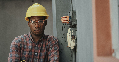 young black man with arms crossed and hardhat next to electrical box